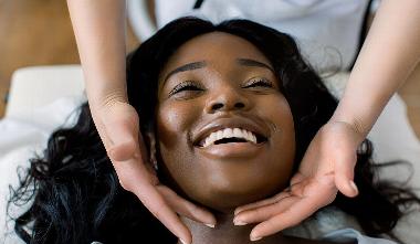 black lady smiling while beautician gestures hands under chin and cheeks from above  